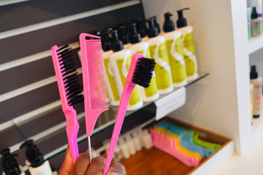 Three pink hair care tools (brush, comb, and brush with black bristles) held in front of a shelf with bottles and bags.